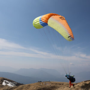 Gleitschirmflieger startet mit orange-gelbem Schirm von einem Berggipfel, im Hintergrund Bergketten