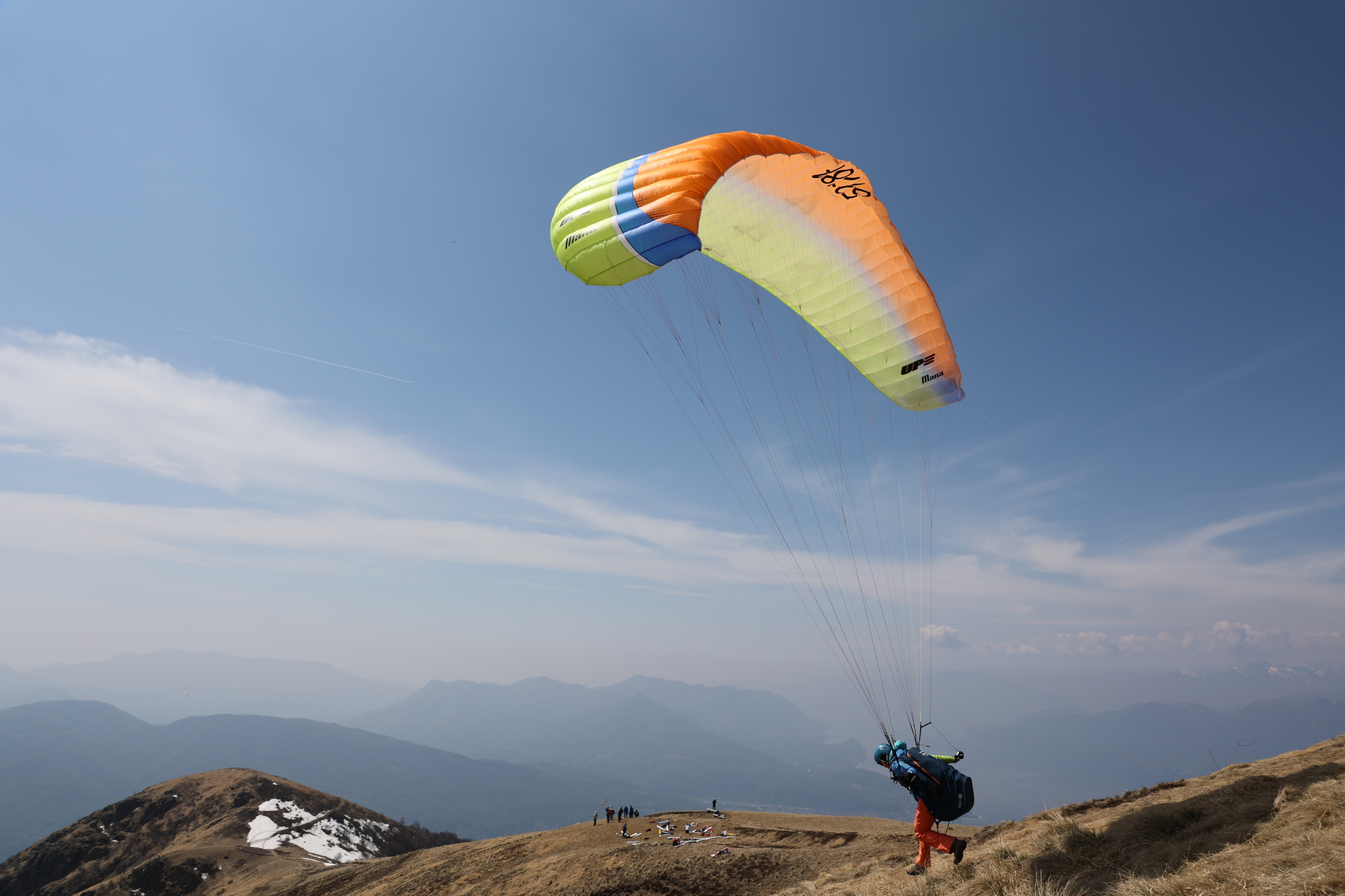 Gleitschirmflieger startet mit orange-gelbem Schirm von einem Berggipfel, im Hintergrund Bergketten
