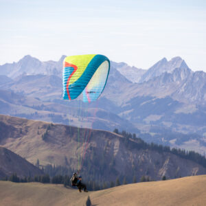 Zwei Personen fliegen im Tandem-Gleitschirm über grüne Berghänge vor einem Alpenpanorama