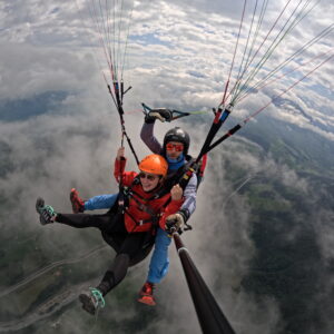 Zwei Personen beim Tandem-Gleitschirmfliegen hoch über Wolken und einer Berglandschaft, die lachend in eine Selfie-Stick-Kamera blicken