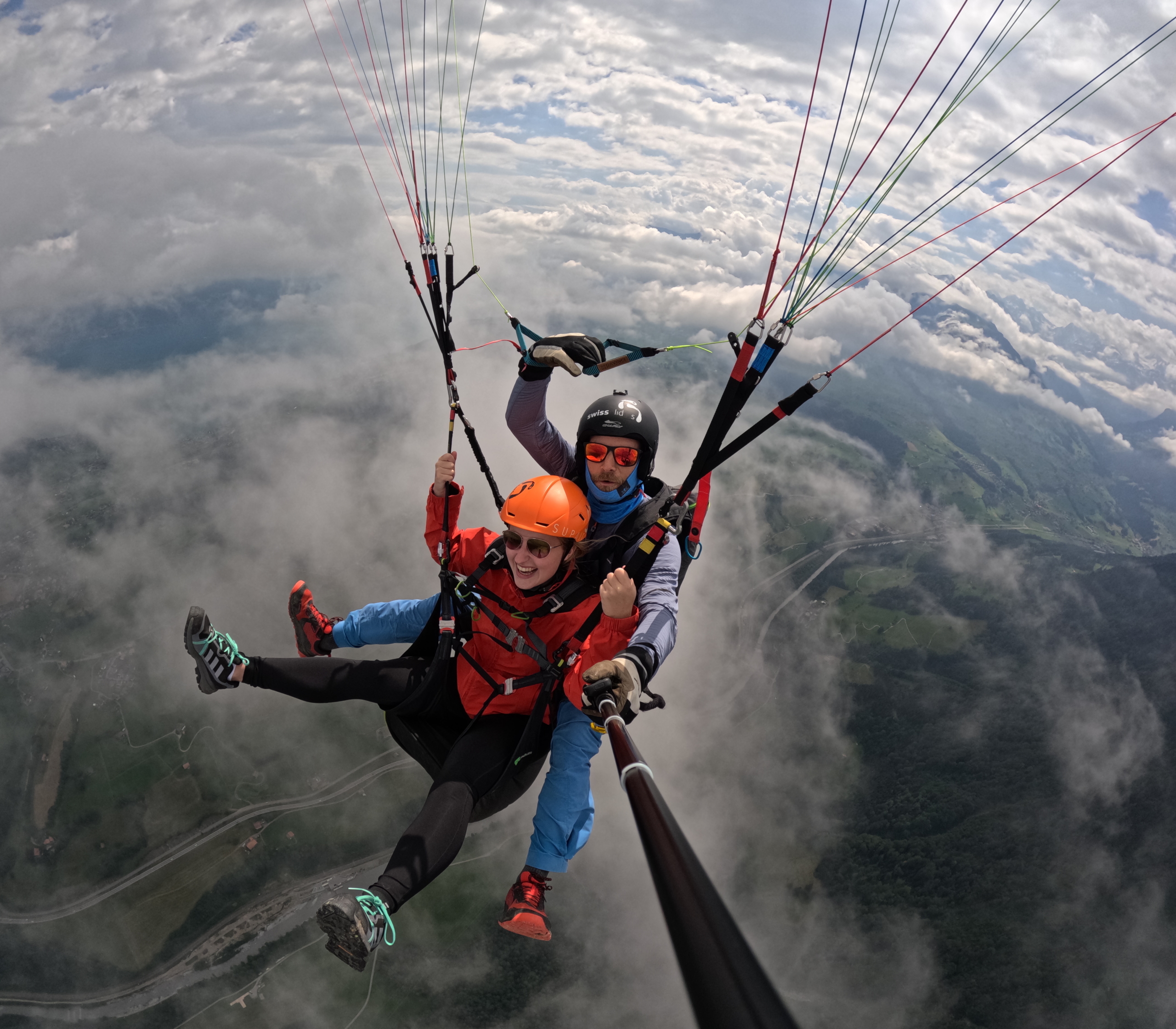 Zwei Personen beim Tandem-Gleitschirmfliegen hoch über Wolken und einer Berglandschaft, die lachend in eine Selfie-Stick-Kamera blicken