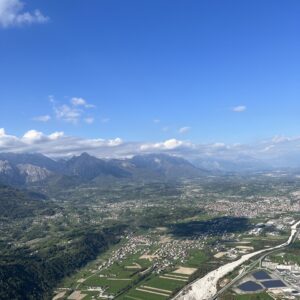 Luftaufnahme eines weiten Alpentals mit Ortschaften, Feldern und einem breiten Flussbett vor Bergkulisse