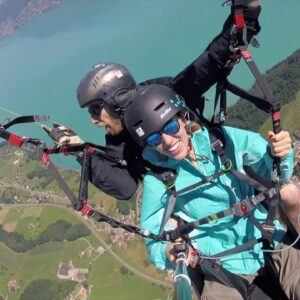 Tandem-Gleitschirmflug hoch über einem türkisfarbenen See und grüner Landschaft, Passagierin lacht in die Kamera