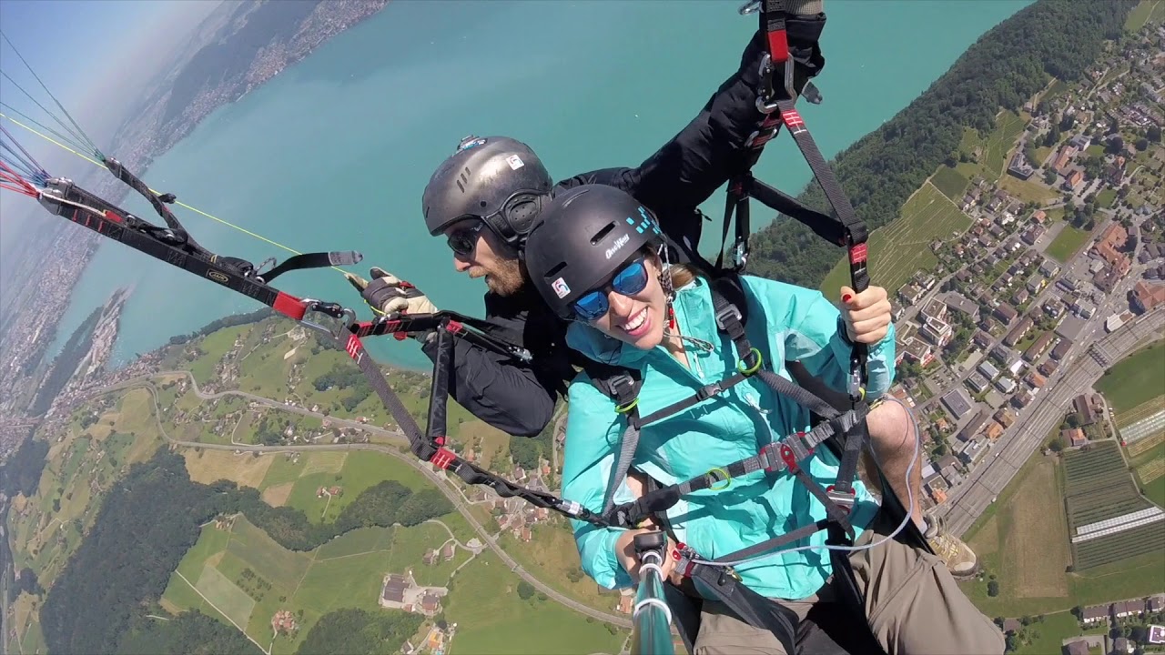 Tandem-Gleitschirmflug hoch über einem türkisfarbenen See und grüner Landschaft, Passagierin lacht in die Kamera