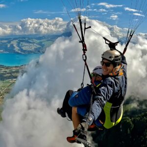 Zwei Personen beim Tandem-Gleitschirmfliegen hoch über Wolken, mit Bergsee und Alpenlandschaft im Hintergrund